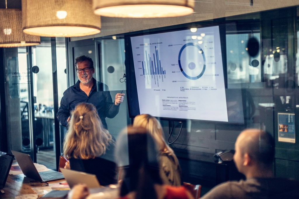 Photo of a mature adult man giving a financial presentation in a modern conference room in an office builfing.